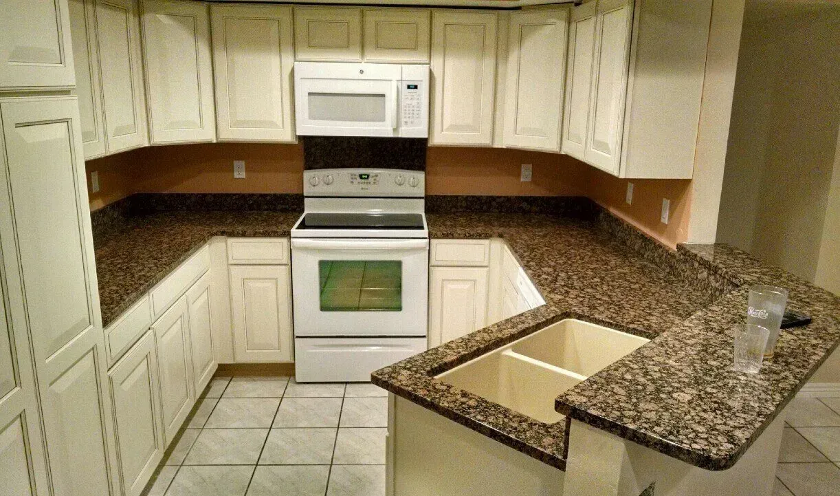 Empty beige kitchen with white cabinets, granite countertops, tiled floor, stove, microwave, and island.