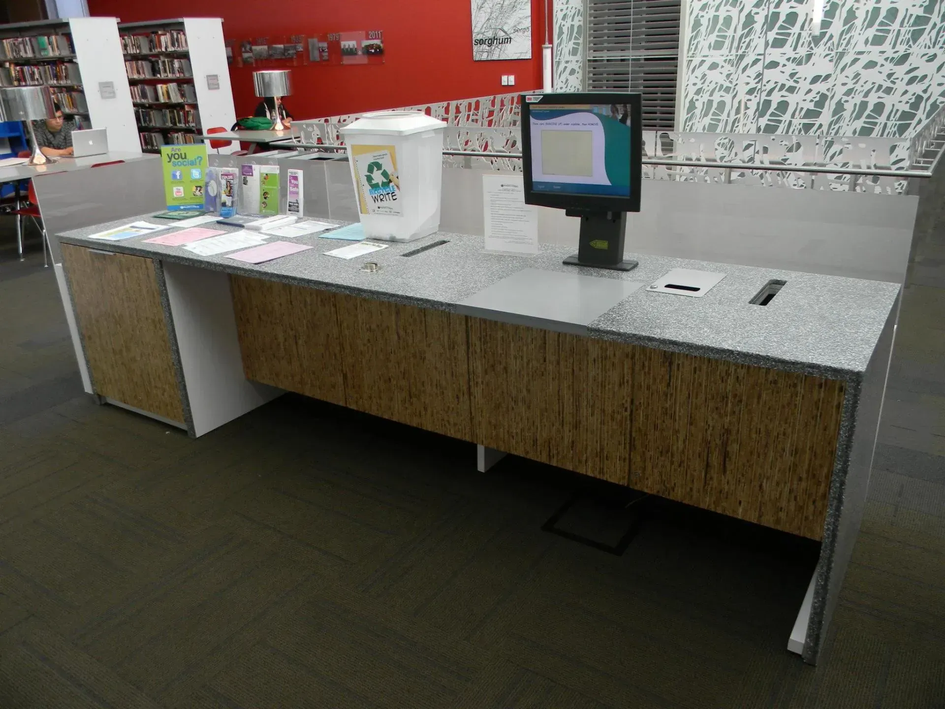 Modern retail checkout counter with a computer monitor, payment terminal, and display brochures in a red-walled store