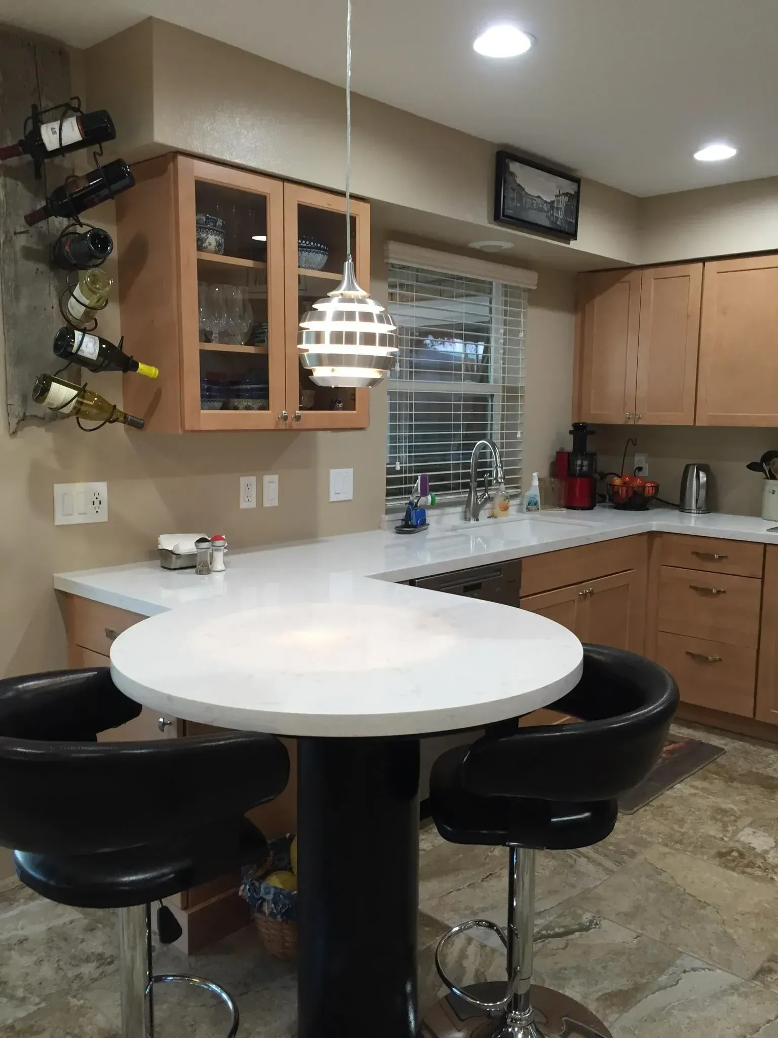 Small kitchen with white round table, black bar stools, and wooden cabinets under warm lights.