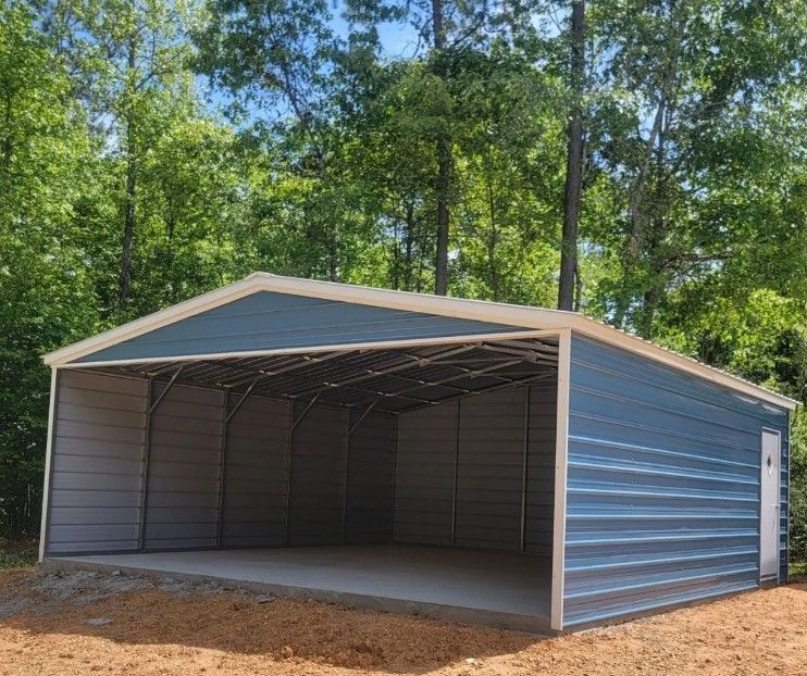 Blue metal carport on a concrete pad, open-sided with a door, set in a wooded area.