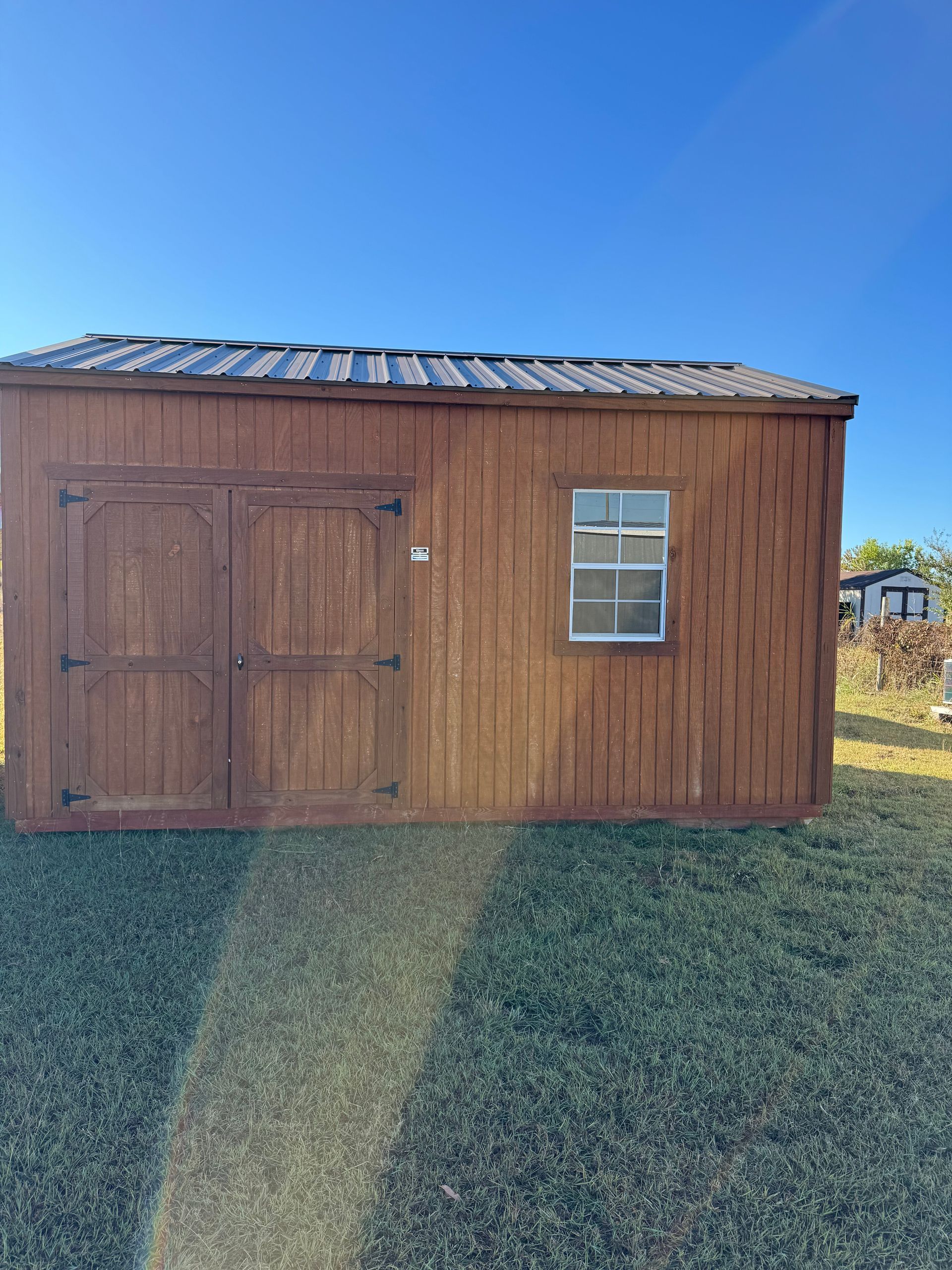 Brown wooden shed with double doors, a small window, and a metal roof on a green lawn.