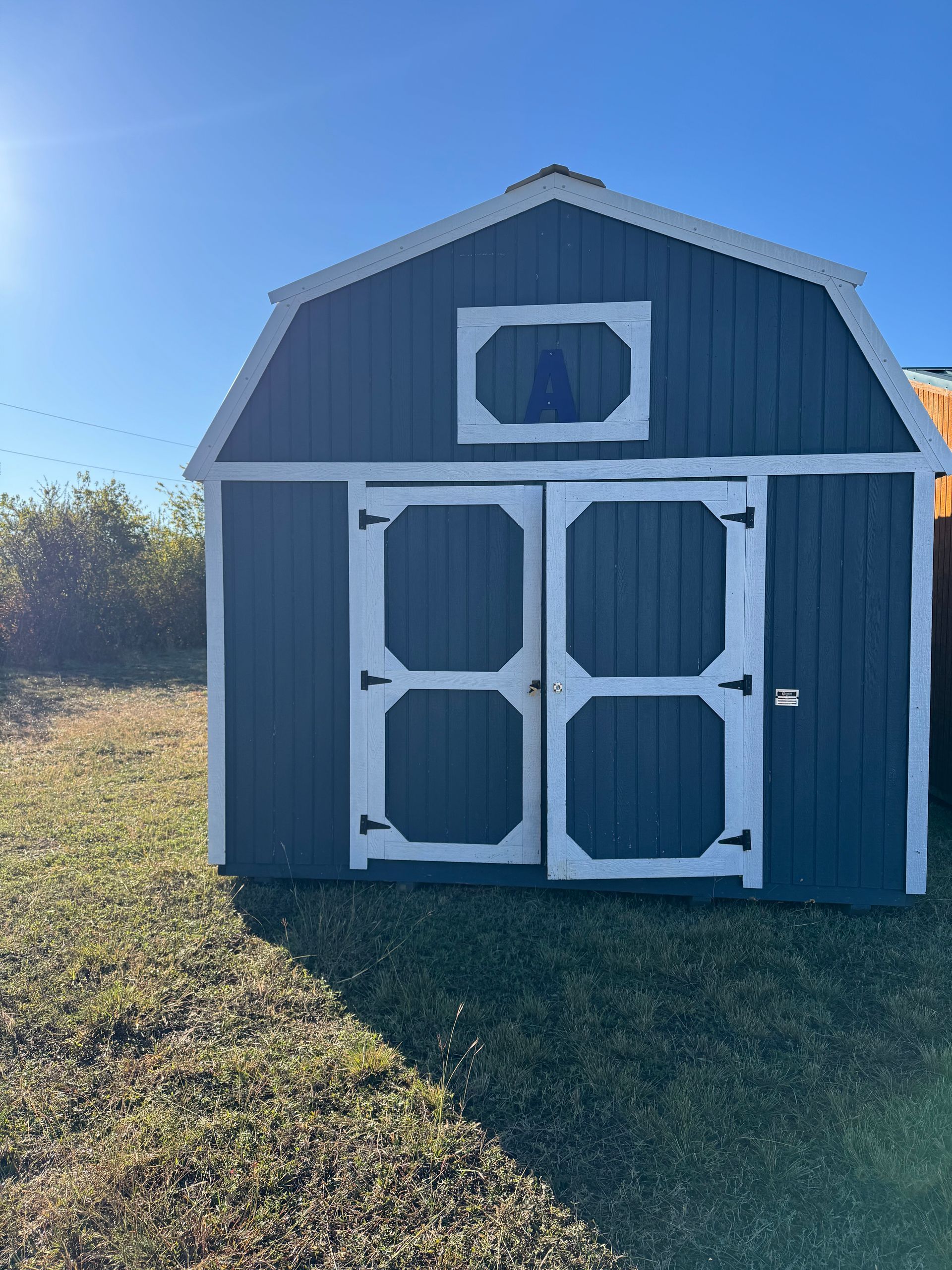 Blue barn-style shed with white trim, double doors, and small upper window, in a grassy area under a blue sky.