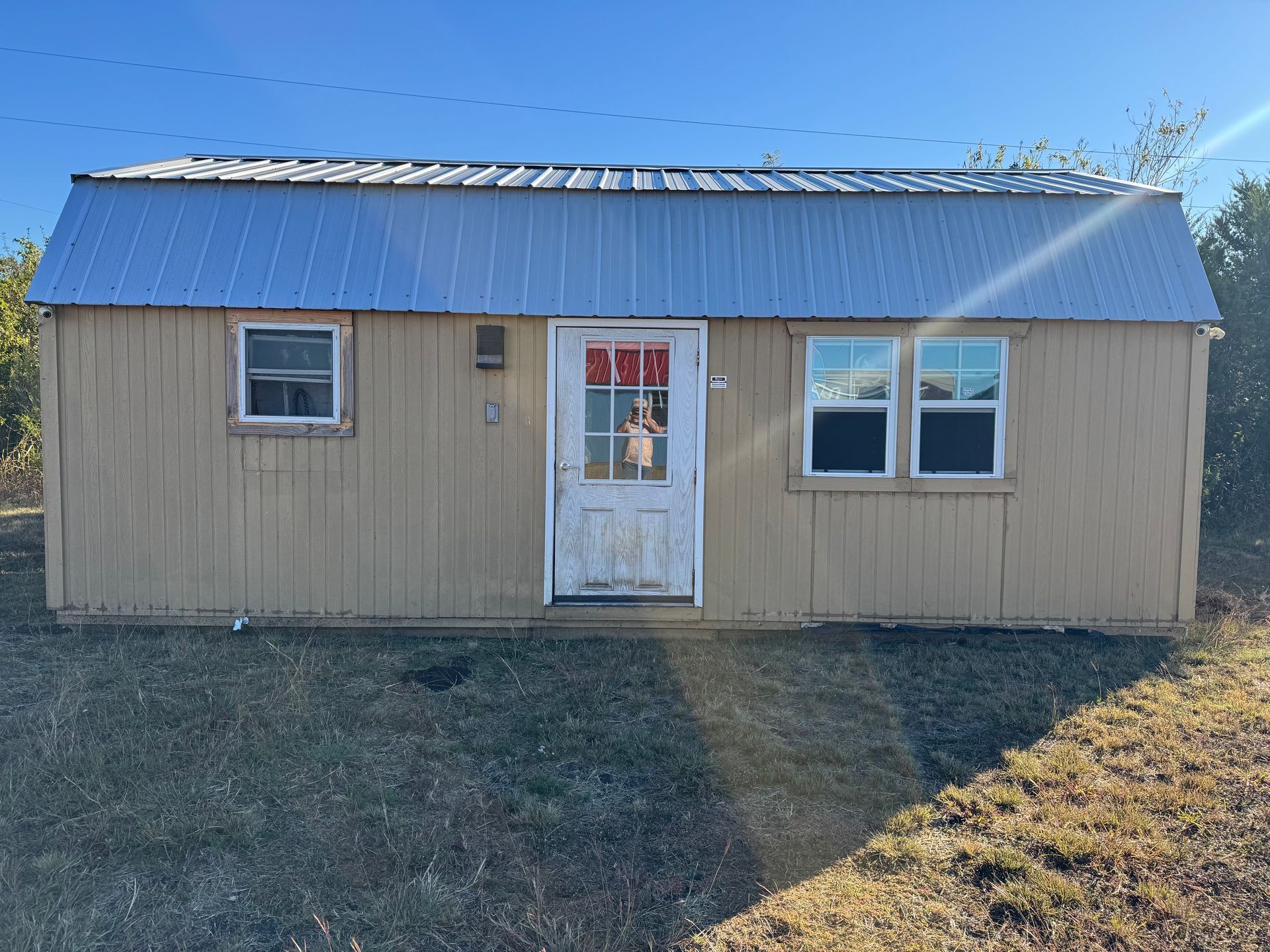 Tan shed with white door and two windows under a metal roof, on a grassy hill.
