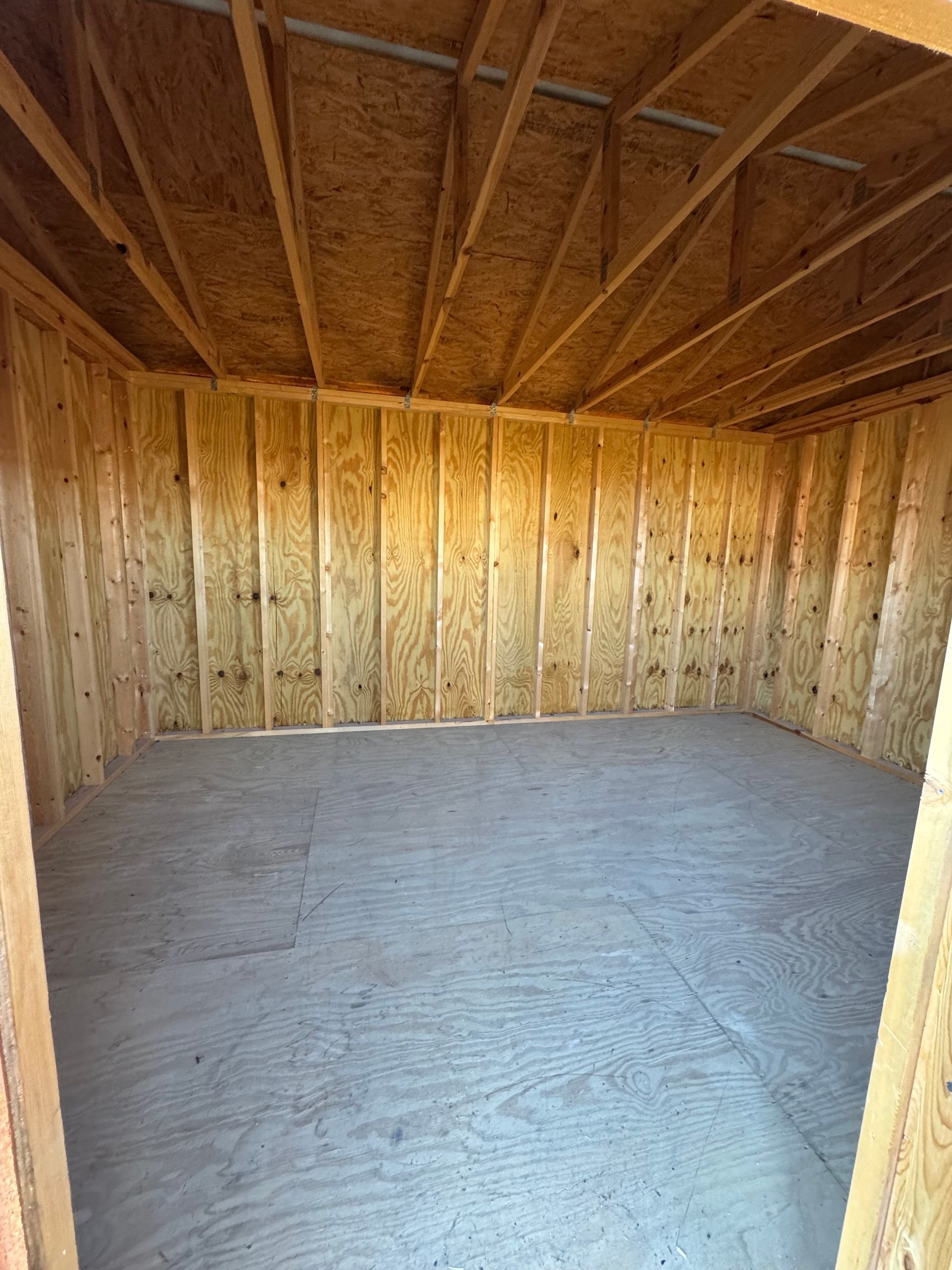 Interior view of a bare wooden room. Walls and ceiling made of exposed wooden studs and plywood. Concrete floor.