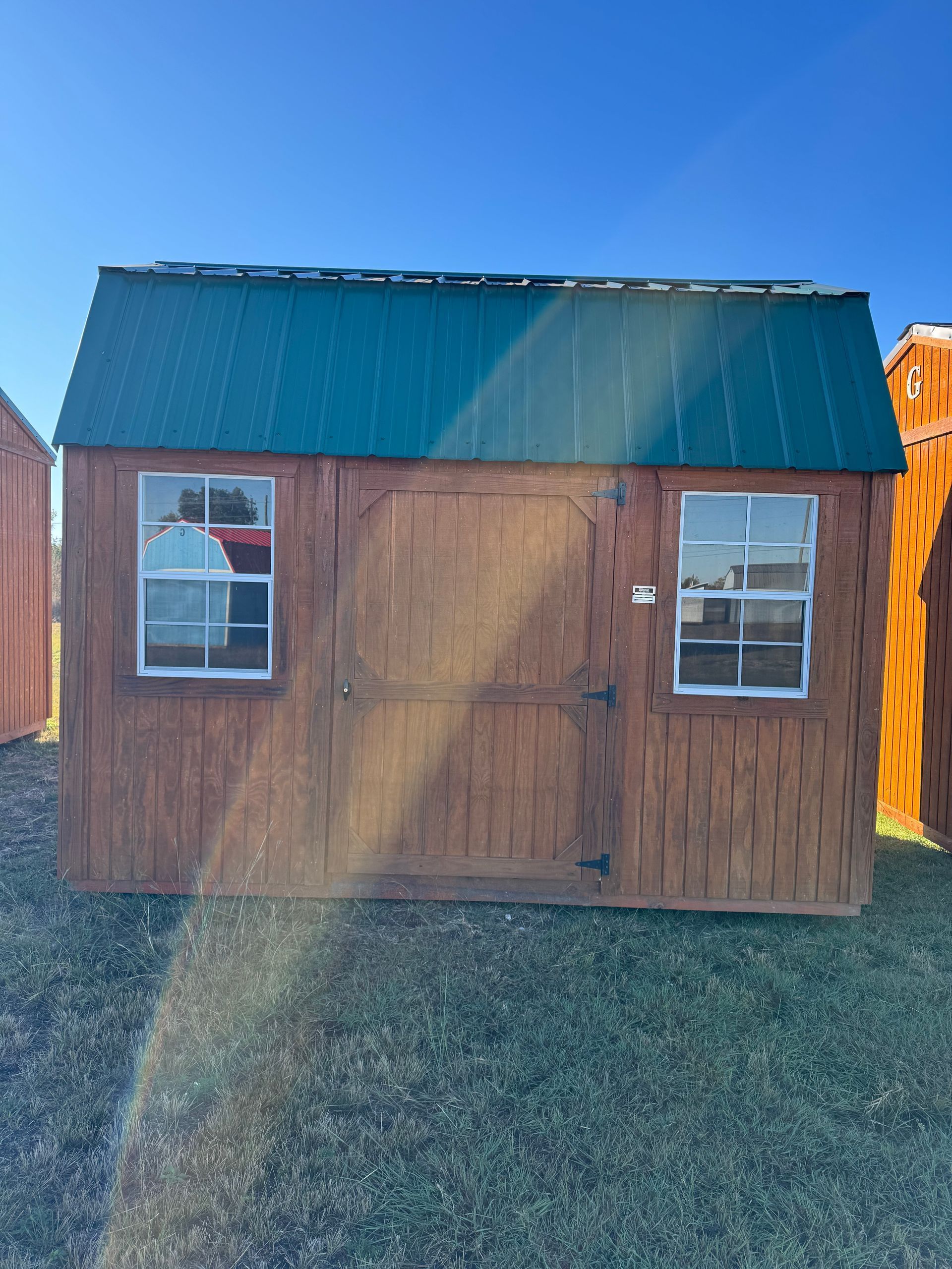 Brown shed with green roof, two windows, and a central door on a grassy field.