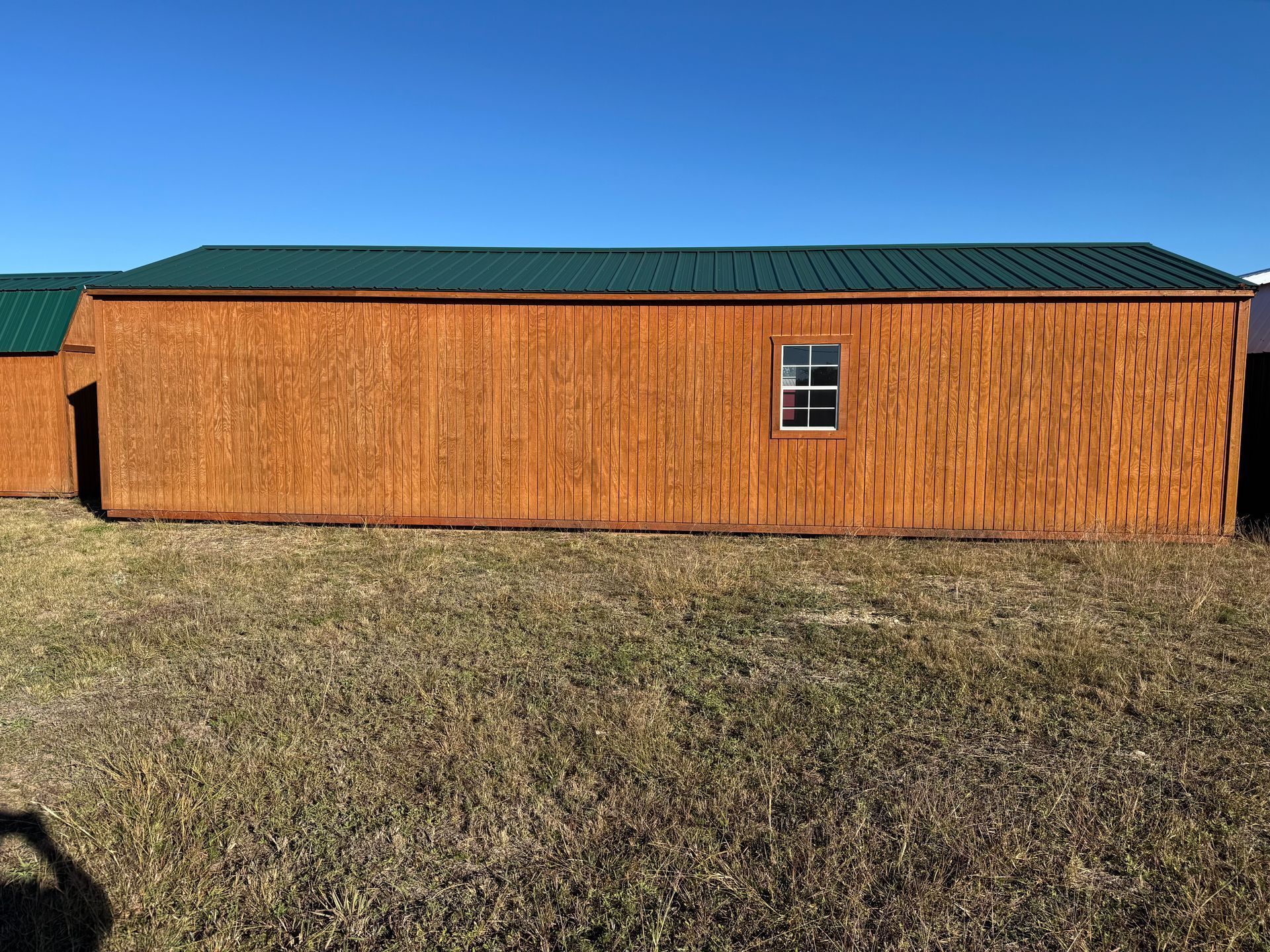 Wooden shed with green metal roof, small window, set in a grassy field under a blue sky.