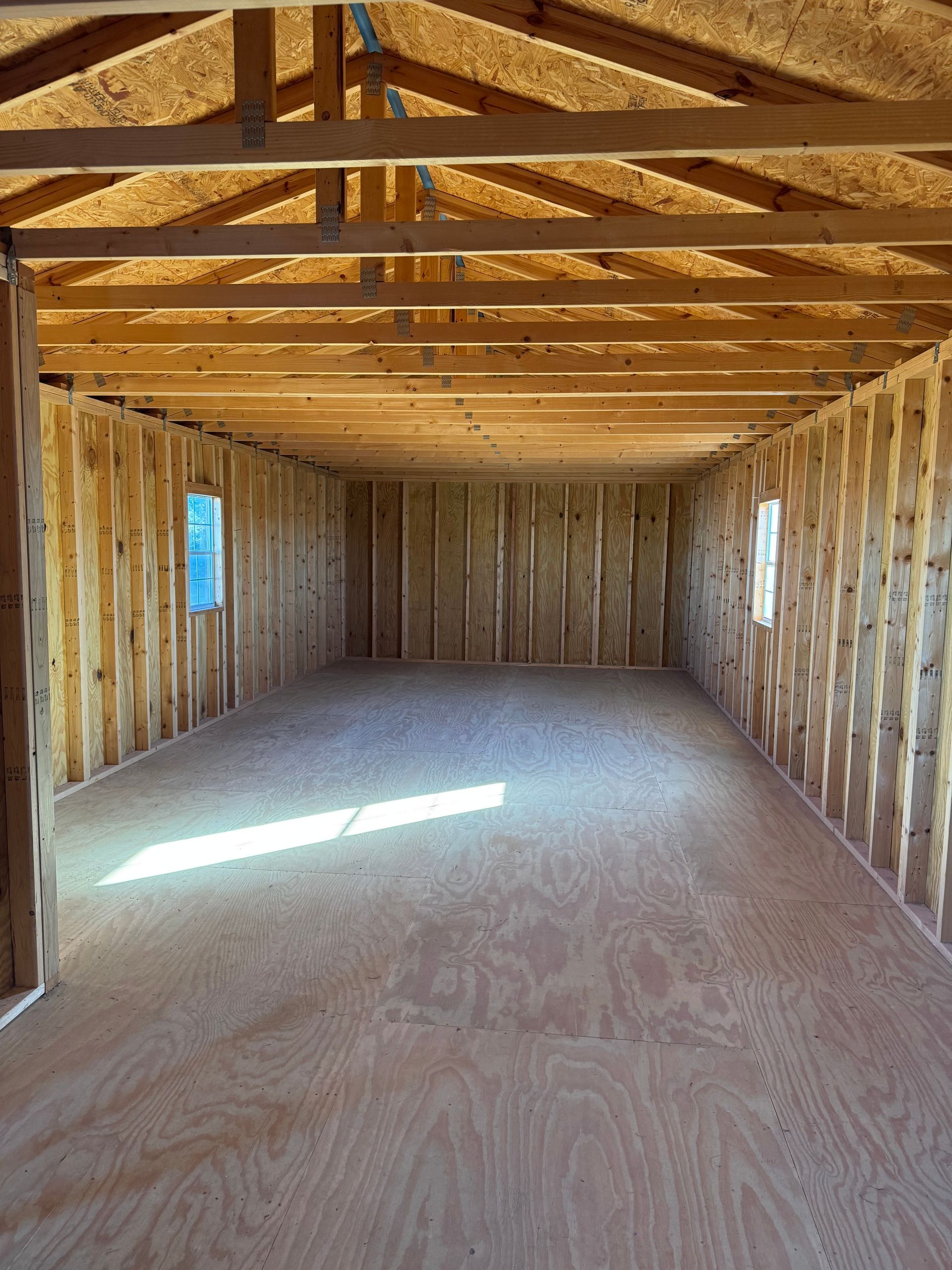 Interior view of a wooden building under construction, with wood flooring, walls, and ceiling framing; two small windows.