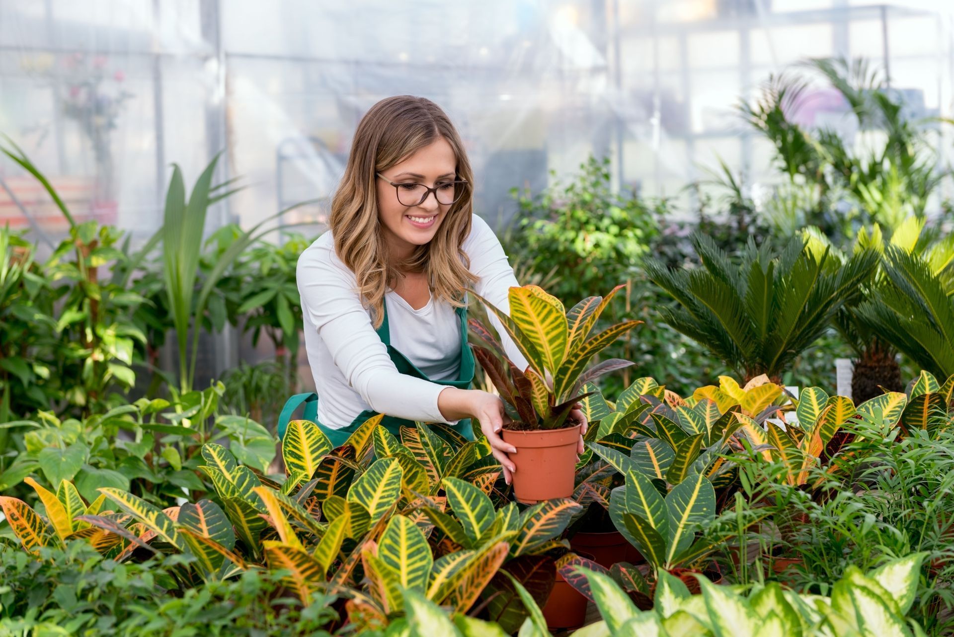 Woman in glasses tending to potted plants in a greenhouse, smiling.
