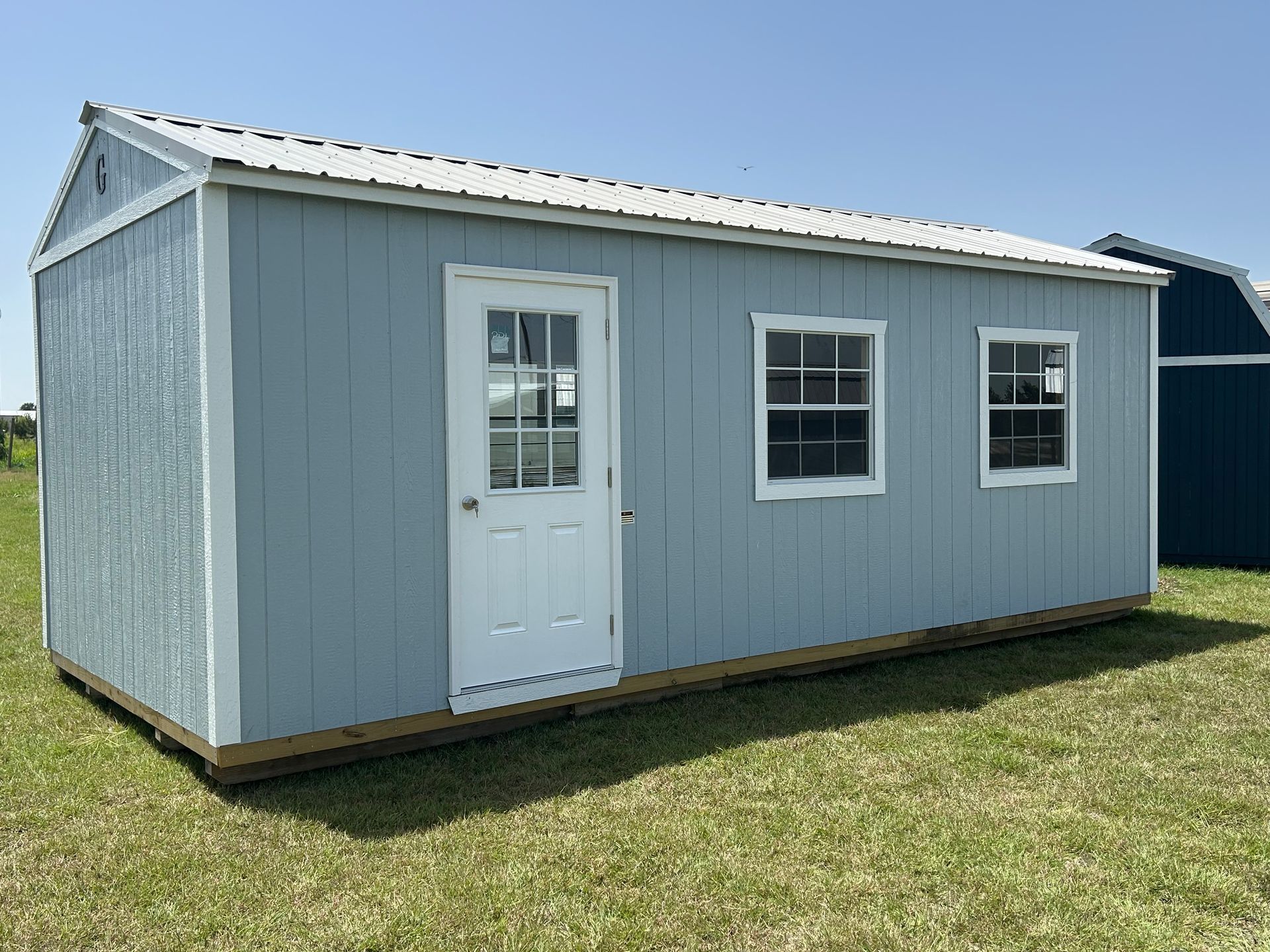 Blue shed with white door, two windows, and metal roof on green grass under a blue sky.