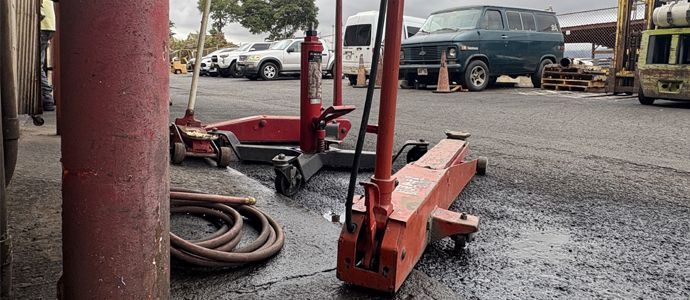 Red hydraulic jacks on wet asphalt, vehicles in the background. Industrial outdoor setting.