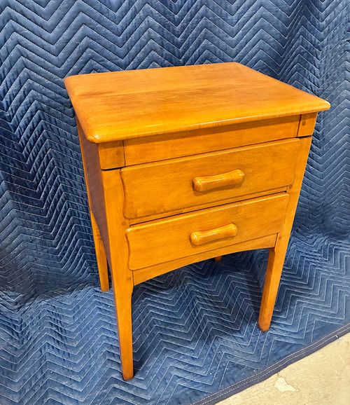 Small wooden nightstand with two drawers and tapered legs on a blue quilted background.
