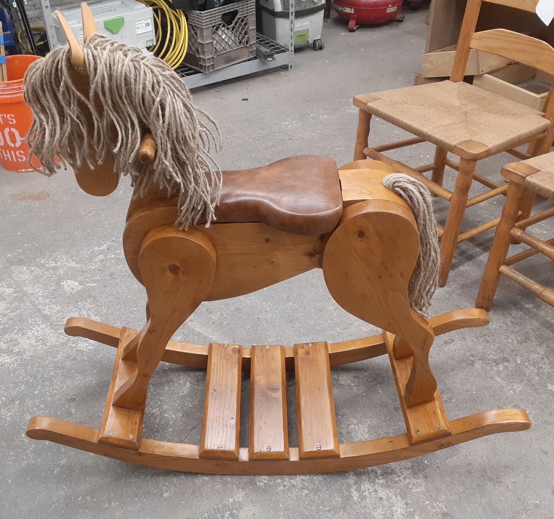 Wooden rocking horse with brown body and rope mane, displayed on a concrete floor in a shop.