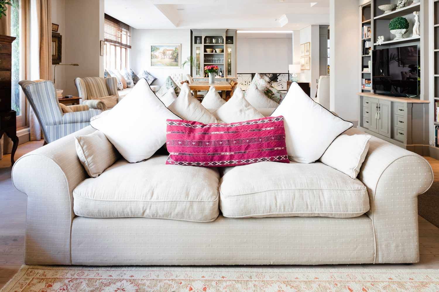 Bright living room with a large cream sofa, white pillows, and a pink accent pillow, centered in a modern space