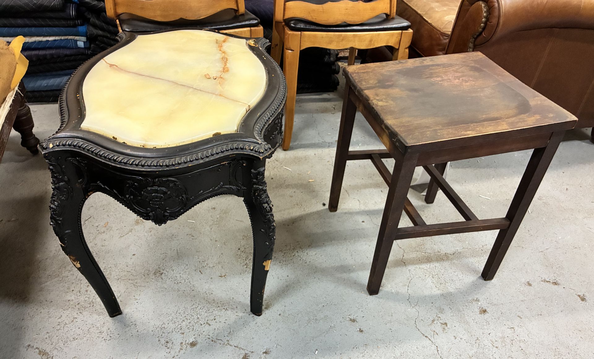 Two small wooden tables, one black with a cream cushion seat and one plain brown stool on a concrete floor.