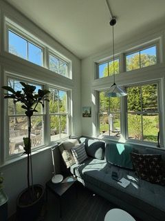 Bright sunroom with large windows, gray couch, hanging light, and potted plant by a lush green view