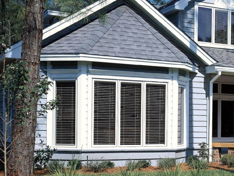 Blue house with a bay window, gray roof, and dark blinds. A tree is in the foreground.