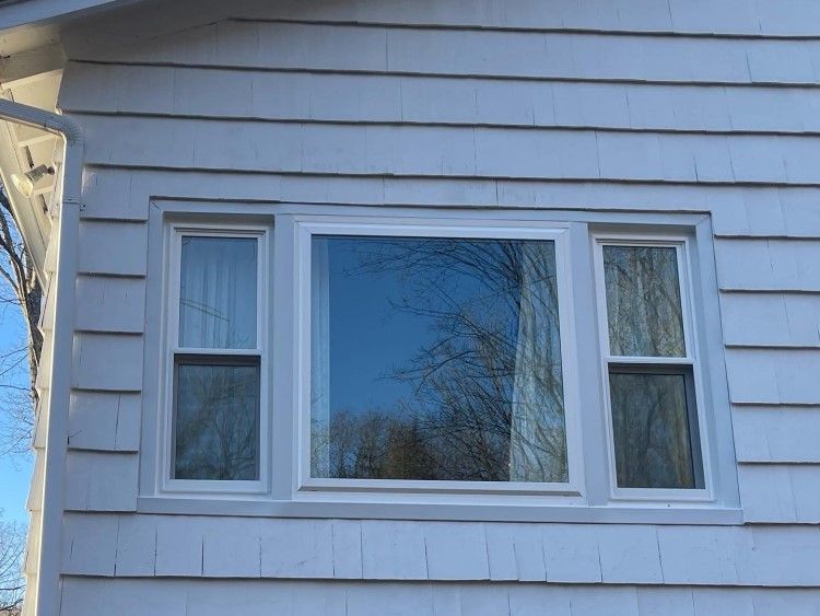 White window on a house with white siding, reflecting trees and a blue sky.