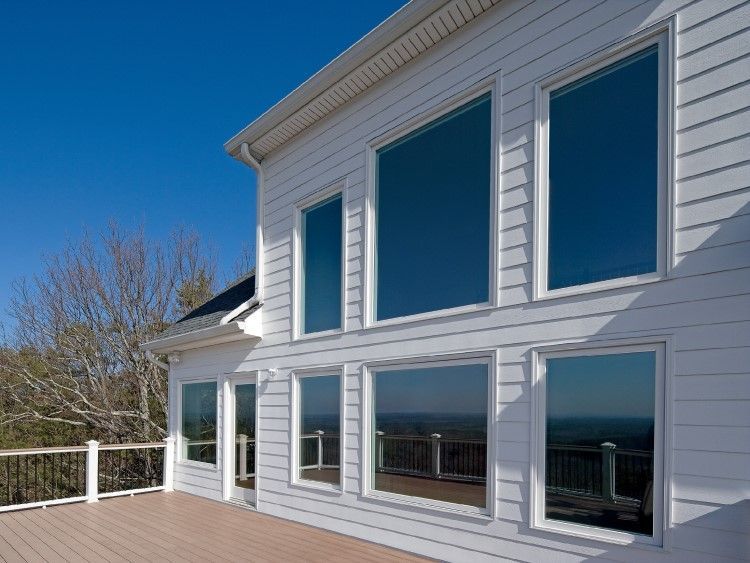 White house with large, reflective windows overlooking a deck and landscape against a bright blue sky.