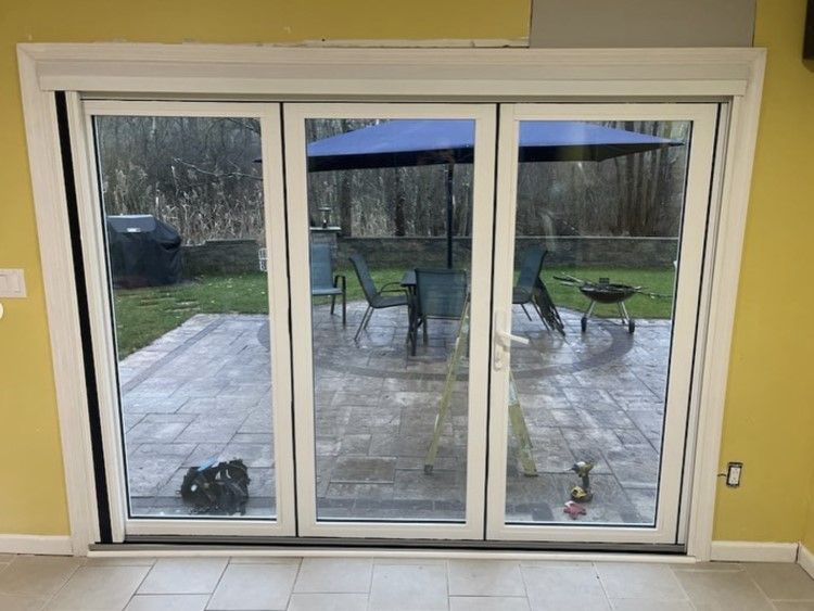 White folding glass doors open to a patio with outdoor furniture and a grill, against a yellow wall.