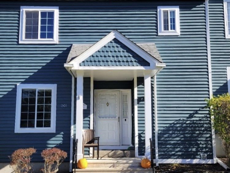 Blue-sided house with a white door, windows, and a small covered entryway; two pumpkins sit on the steps.
