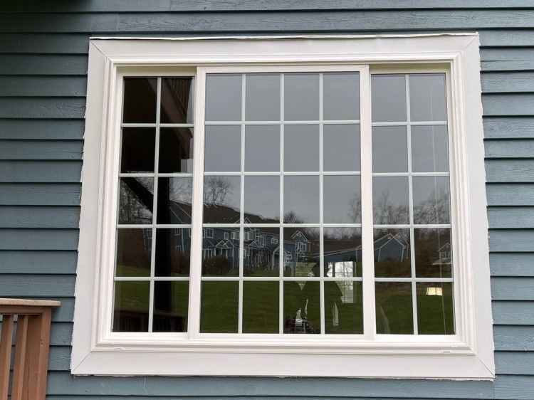 White-framed sliding window with multiple panes, reflecting a house and trees, set in blue siding.