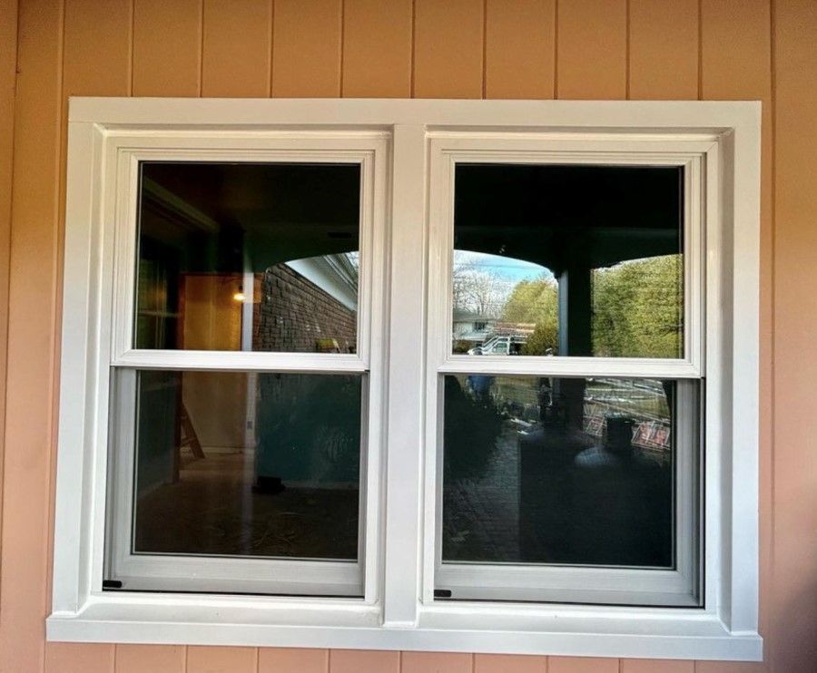 White-framed double-hung windows set in an exterior wall with light orange siding; reflecting views of outdoors.