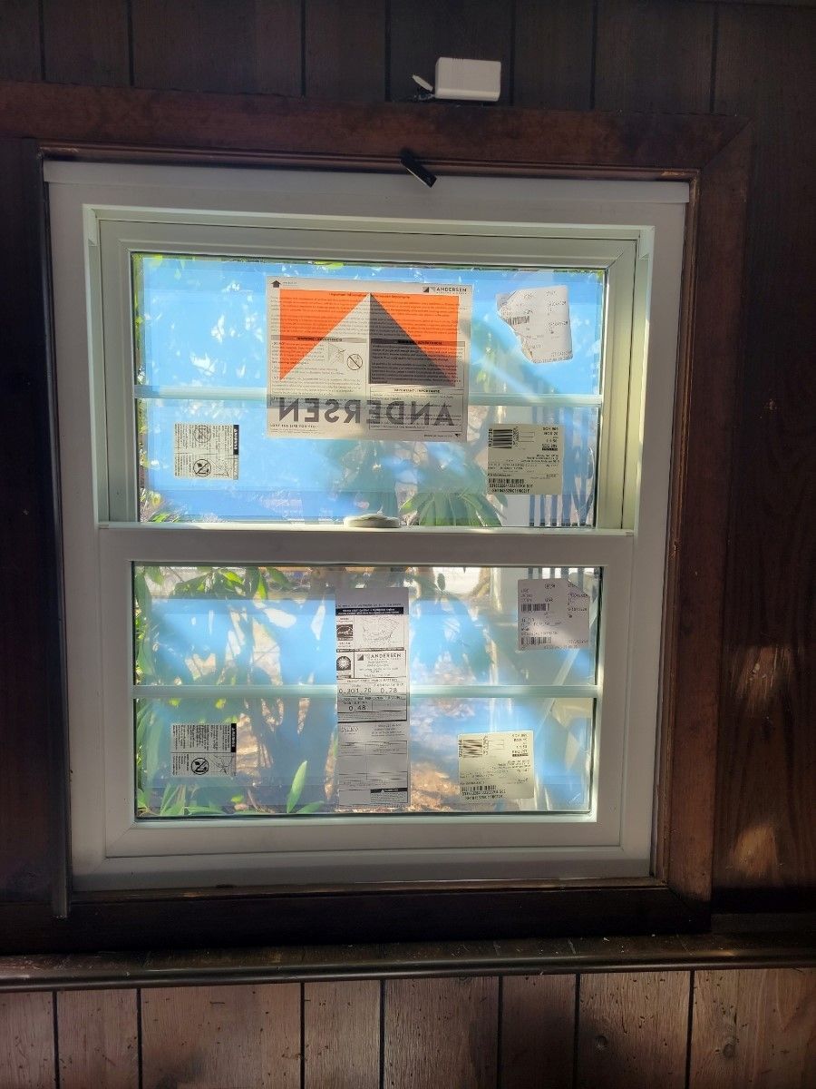 Newly installed white window with protective blue wrapping and a white frame, inside a wood-paneled wall.