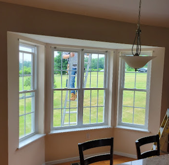Person on ladder cleaning a three-pane bay window. Green outdoors visible. Beige walls and white window frames.