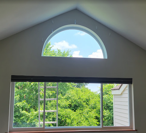 Two windows: a half-circle above a rectangular one, with shades. Green trees and blue sky are visible through the windows.