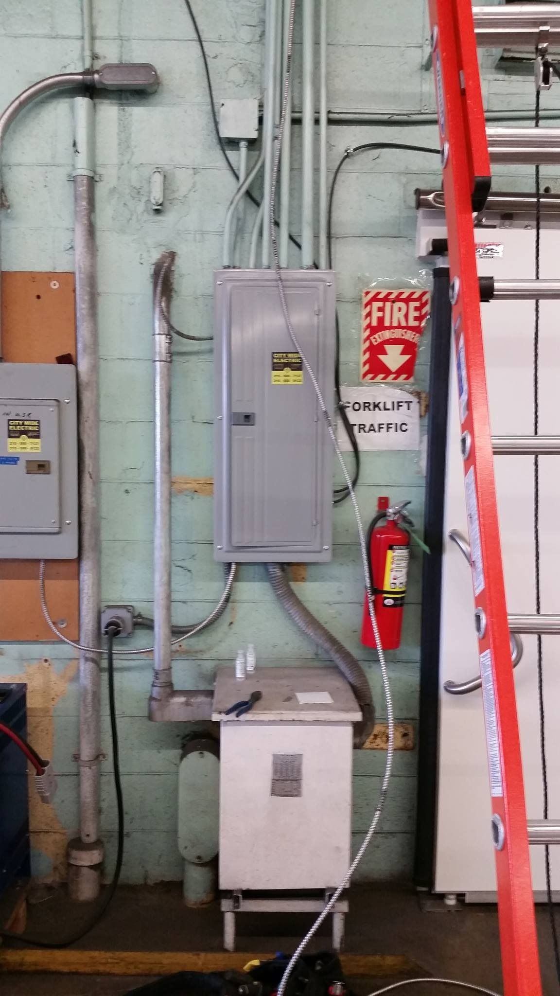 Electrical panel and fire extinguisher on industrial wall. Red ladder leans nearby.