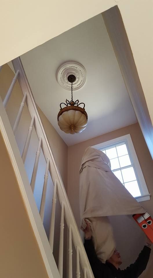 Person on ladder, reaching for light fixture in stairwell. White walls, banister, and ceiling.