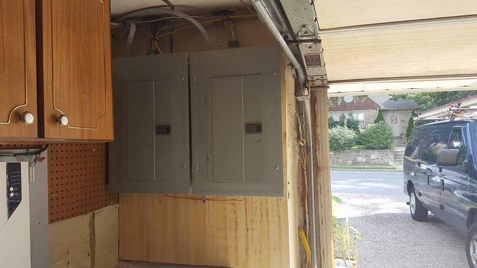 Two gray electrical panels mounted on a wooden wall under a carport, a blue van is visible.