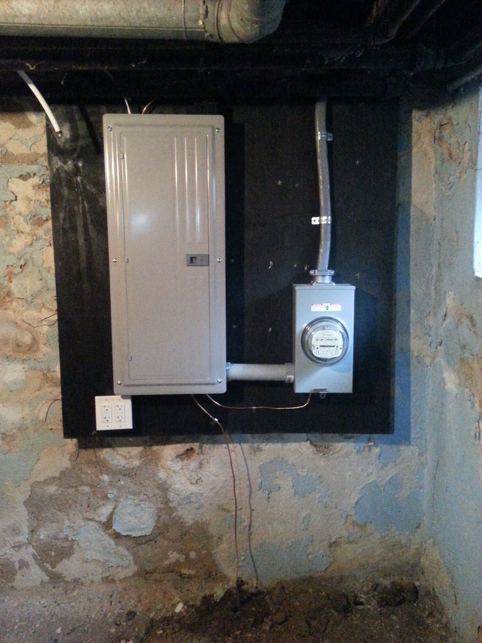 Electrical panel and meter on a black backing, mounted on a concrete basement wall.