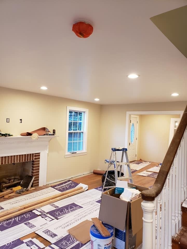 Interior room undergoing renovation; tan walls, new flooring, fireplace, staircase, ladder, and supplies are present.
