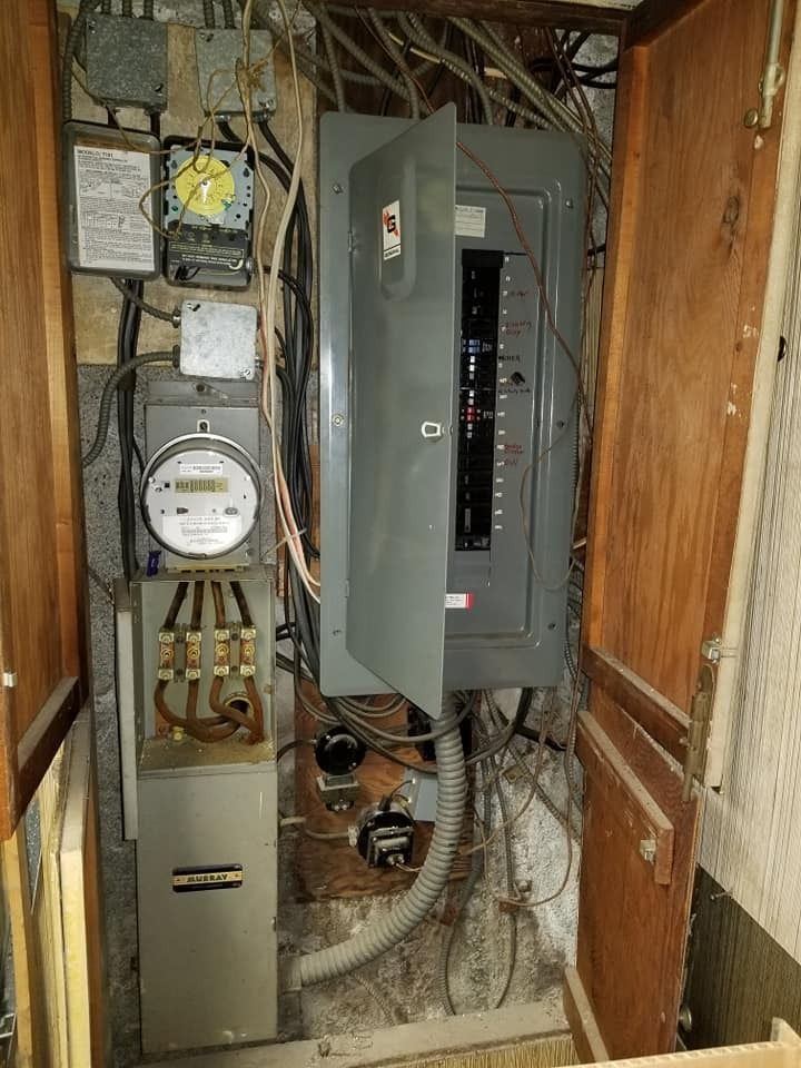 Electrical panel with open door, surrounded by wiring and wooden panels in a confined space.