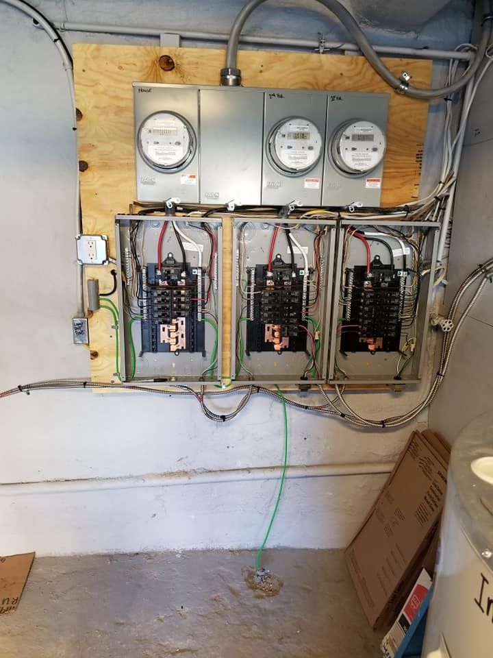 Electrical panel with three meters and circuit breaker boxes mounted on plywood in a concrete room.