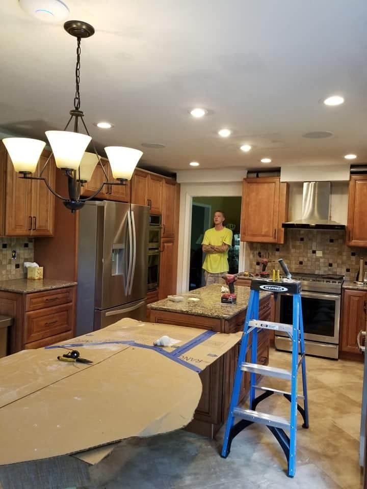 Kitchen renovation in progress: A person stands in a doorway, ladder in the foreground. Island with cardboard on top, appliances.