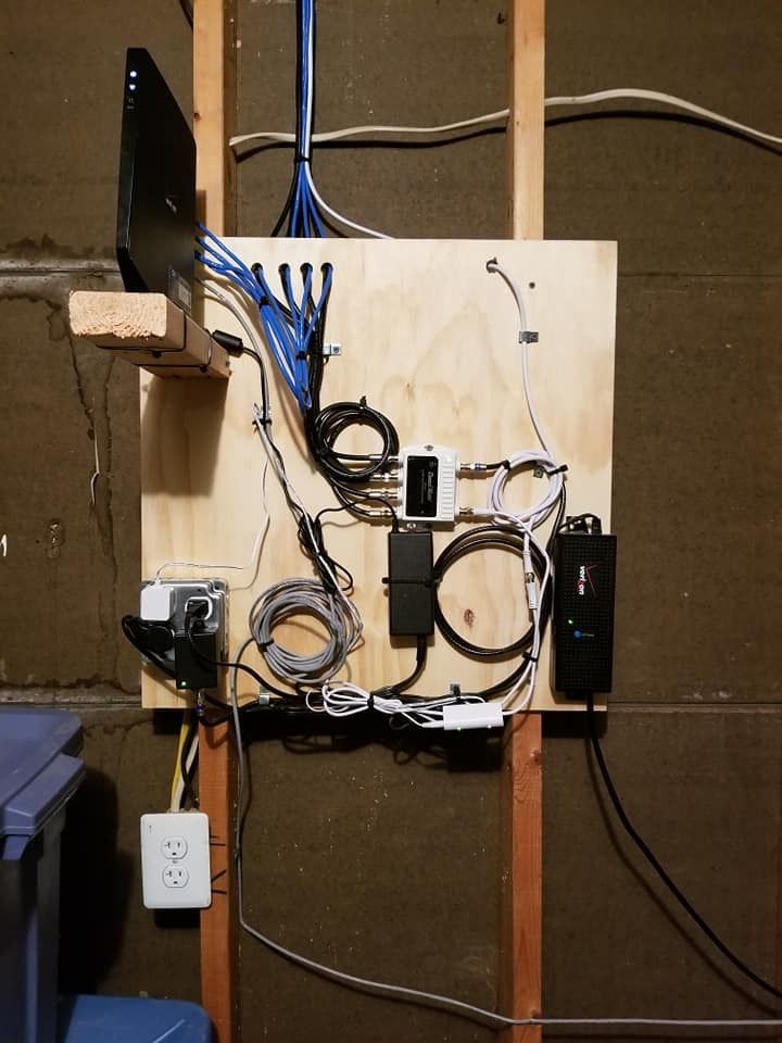 Network wiring panel with various cables and devices mounted on plywood, attached to studs in a wall.
