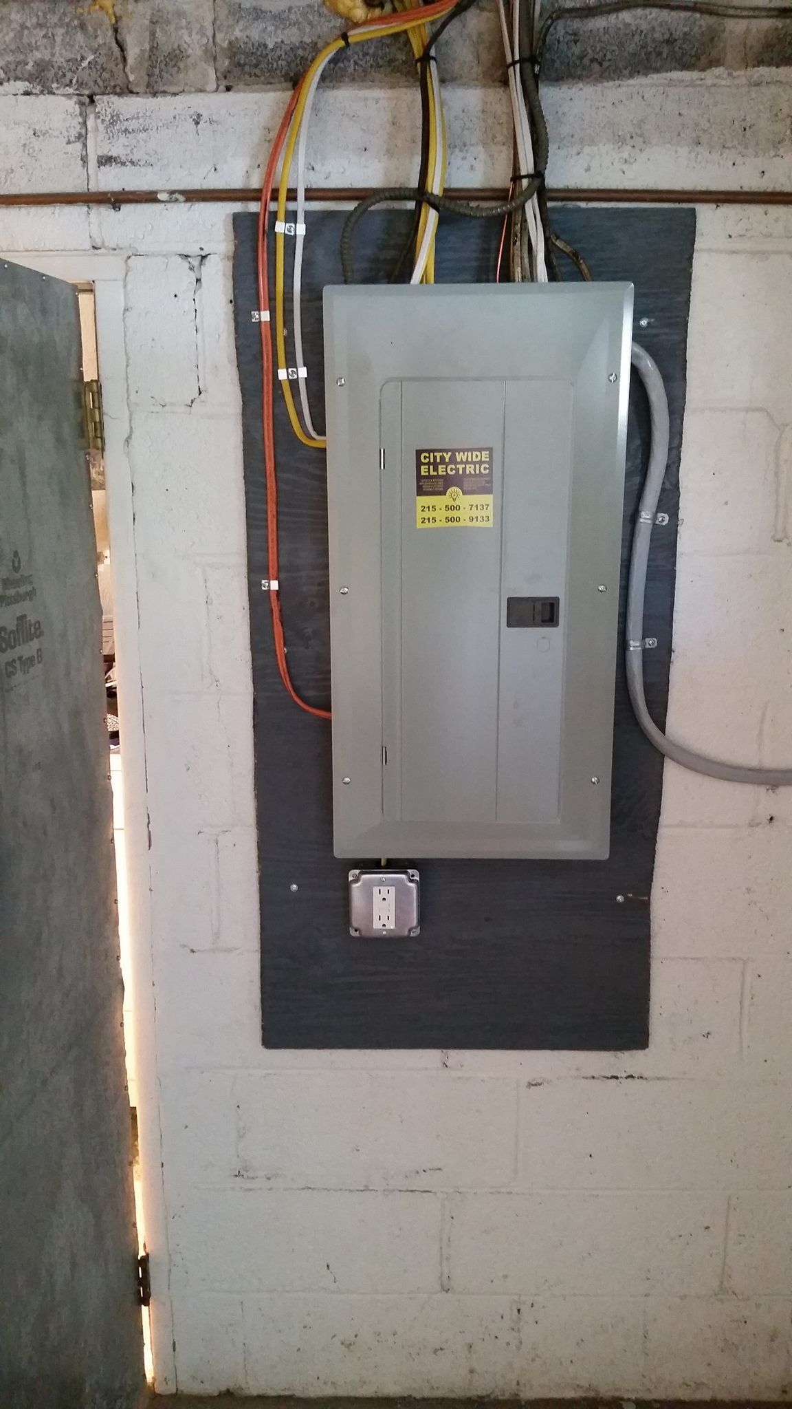 Electrical panel with conduit and wires mounted on a wooden board on a white cinderblock wall. An outlet is below.