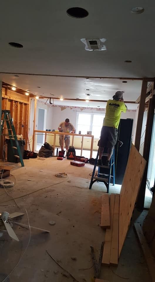 Construction workers on ladders inside a room undergoing renovation. Exposed walls, ceiling fixtures, tools visible.