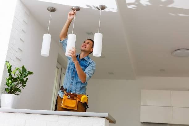 Man installing hanging lights in a modern kitchen. He's standing on a counter, wearing a tool belt, and reaching upwards.