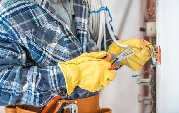 Electrician in plaid shirt and gloves, using pliers on wiring near an outlet.