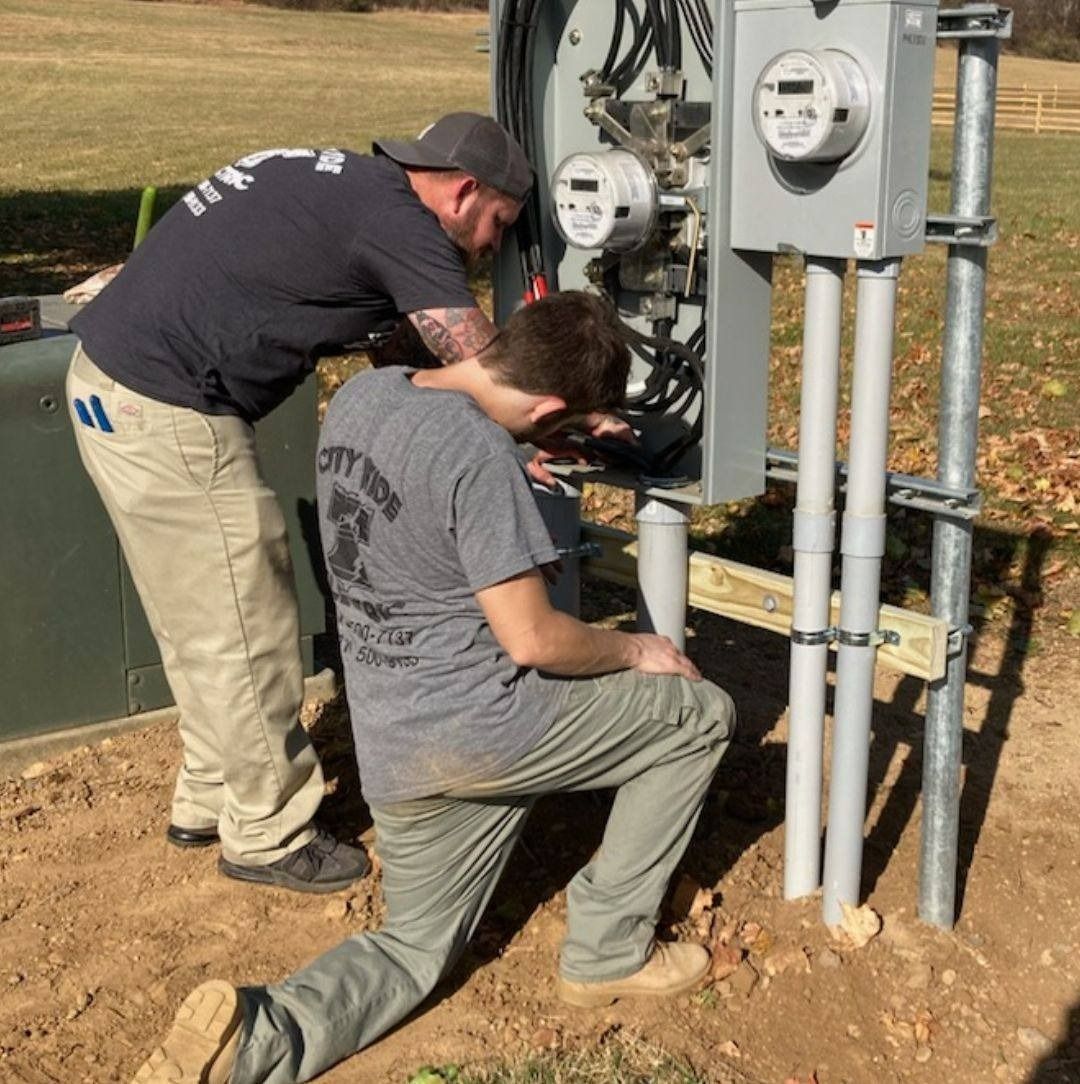 Two electricians working on electrical meters outdoors. One kneels while the other observes.