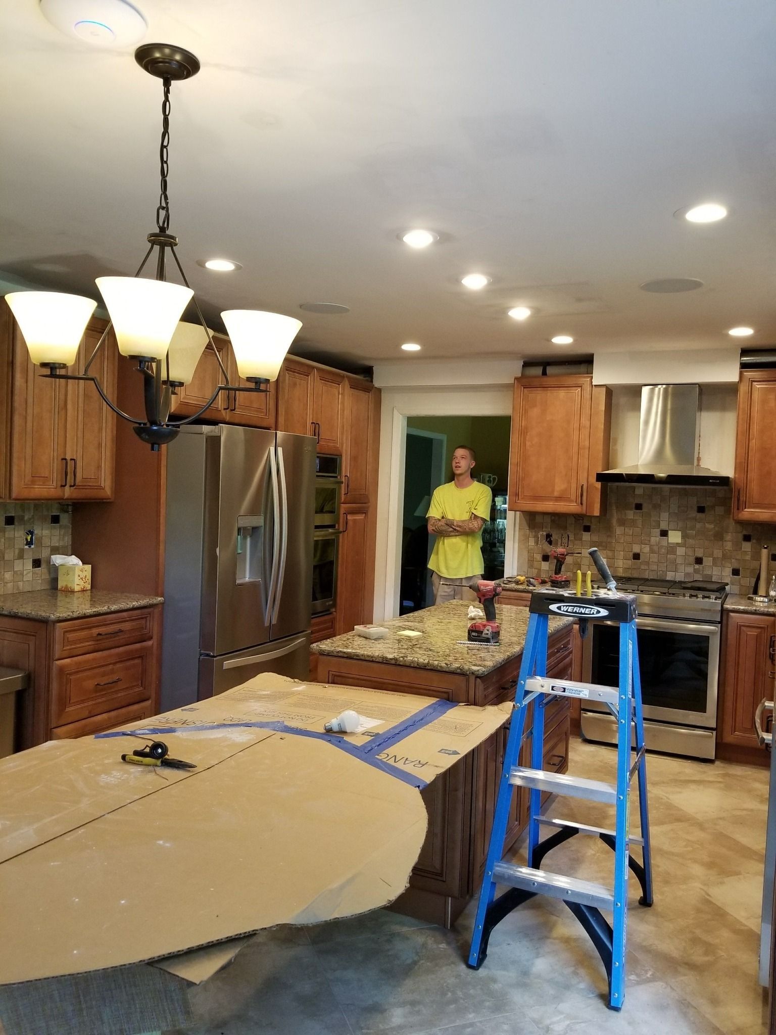 Kitchen renovation in progress: Person in yellow shirt, island with cardboard, ladder, cabinets, chandelier.