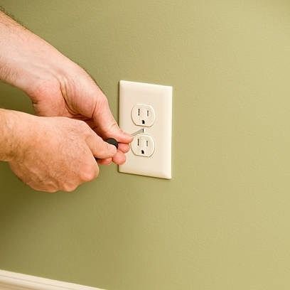Hands installing an electrical outlet on a green wall with a screwdriver.