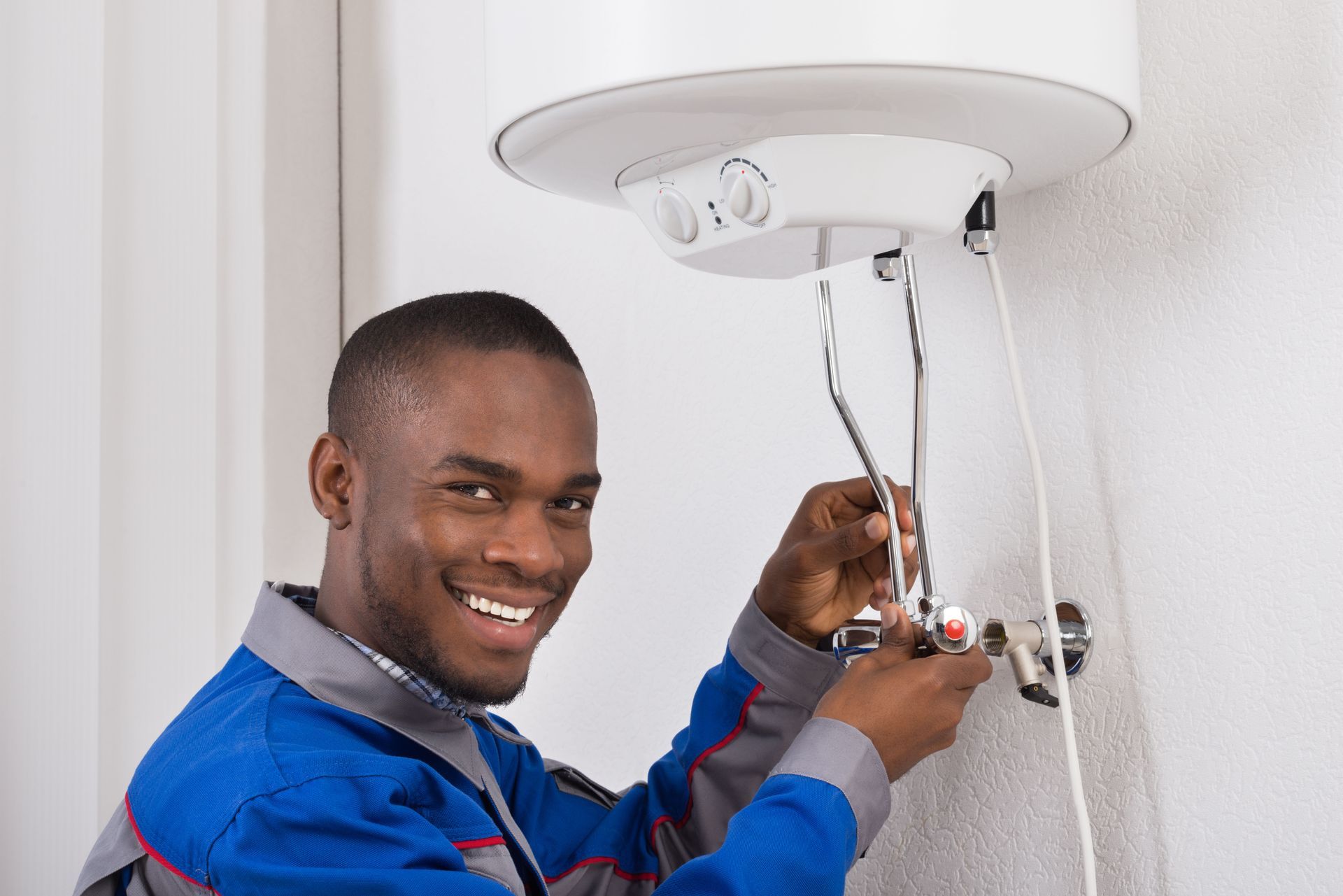 Plumber in blue uniform smiling while working on a water heater, indoors.