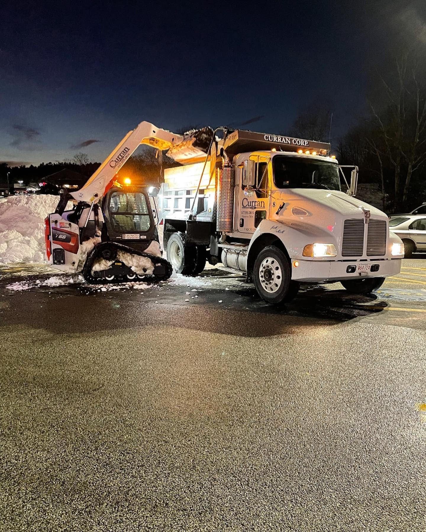 A white skid steer loader lifts a bucket of snow into the bed of a white dump truck parked in a lit, snowy parking lot.