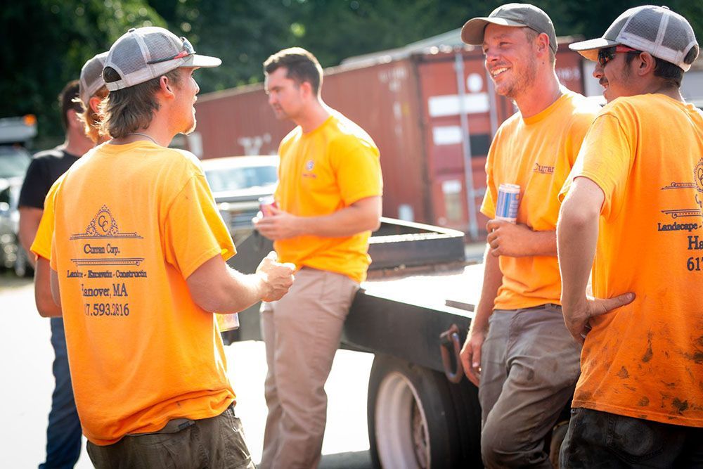 The experts of Curran Corp Landscape & Excavation in orange shirts and caps are talking beside a truck at a construction site.