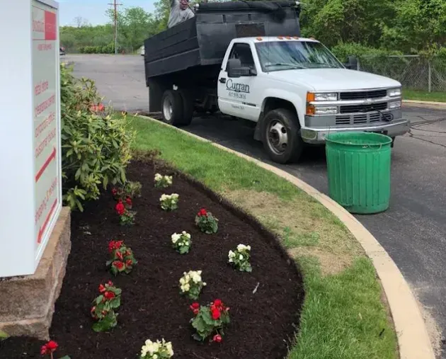 A white dump truck is parked near a mulched garden bed with small red and white flowers at a commercial property.
