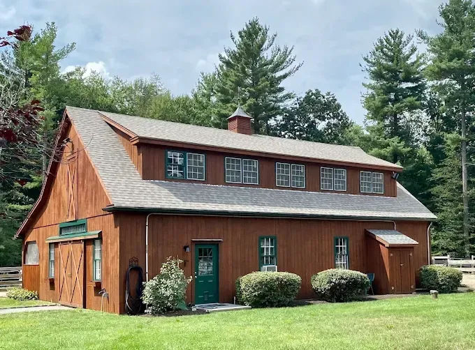 A brown barn with a dormer, a green door, and small square windows, surrounded by green lawn and tall trees.